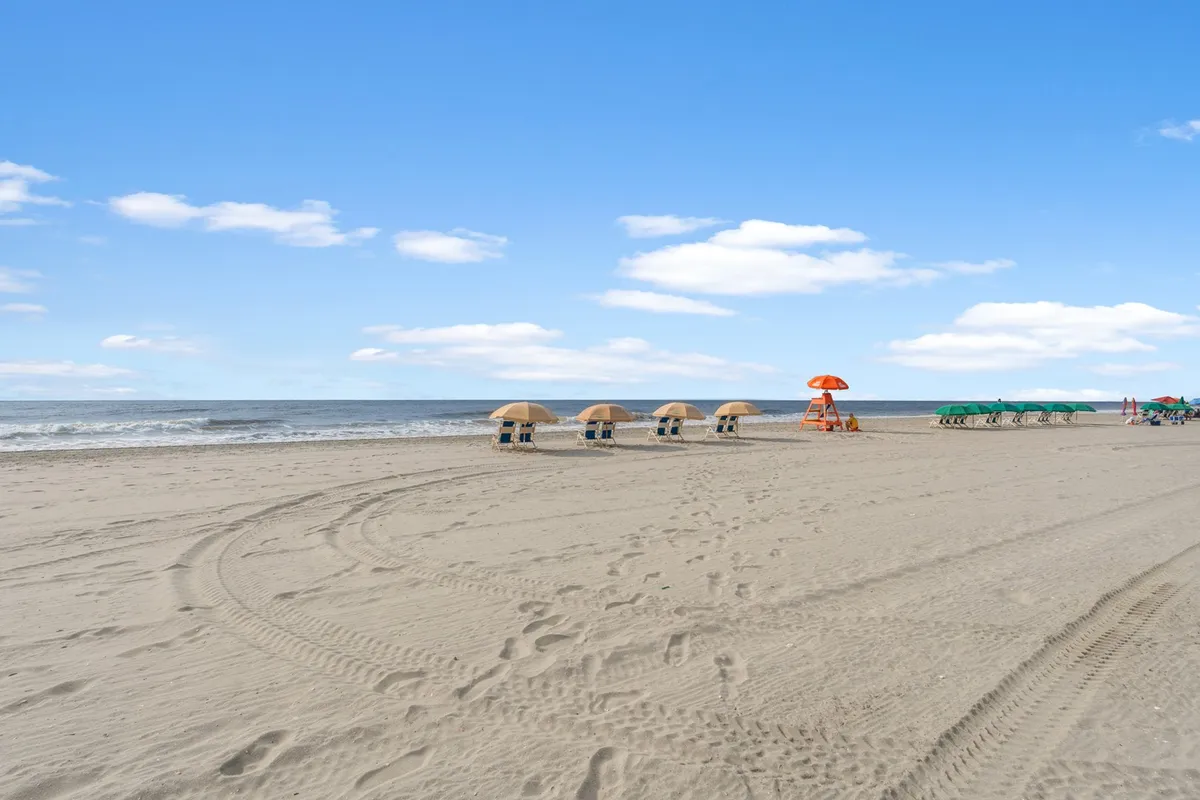 A pristine beach scene featuring beach umbrellas, a lifeguard stand, rows of beach chairs facing gentle ocean waves, and a wide stretch of sand. The mood is cheerful and carefree, evoking the classic Myrtle Beach oceanfront experience.