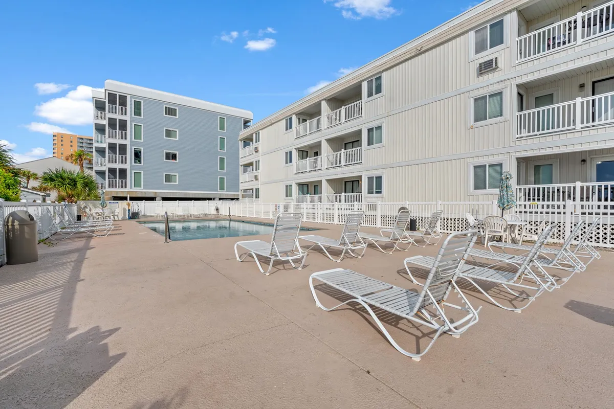 A sparkling swimming pool surrounded by lounge chairs and palm trees sits adjacent to the building, creating an inviting resort-like atmosphere. The area is set up for both relaxing and socializing, with ample space for sunbathing and gathering in groups.