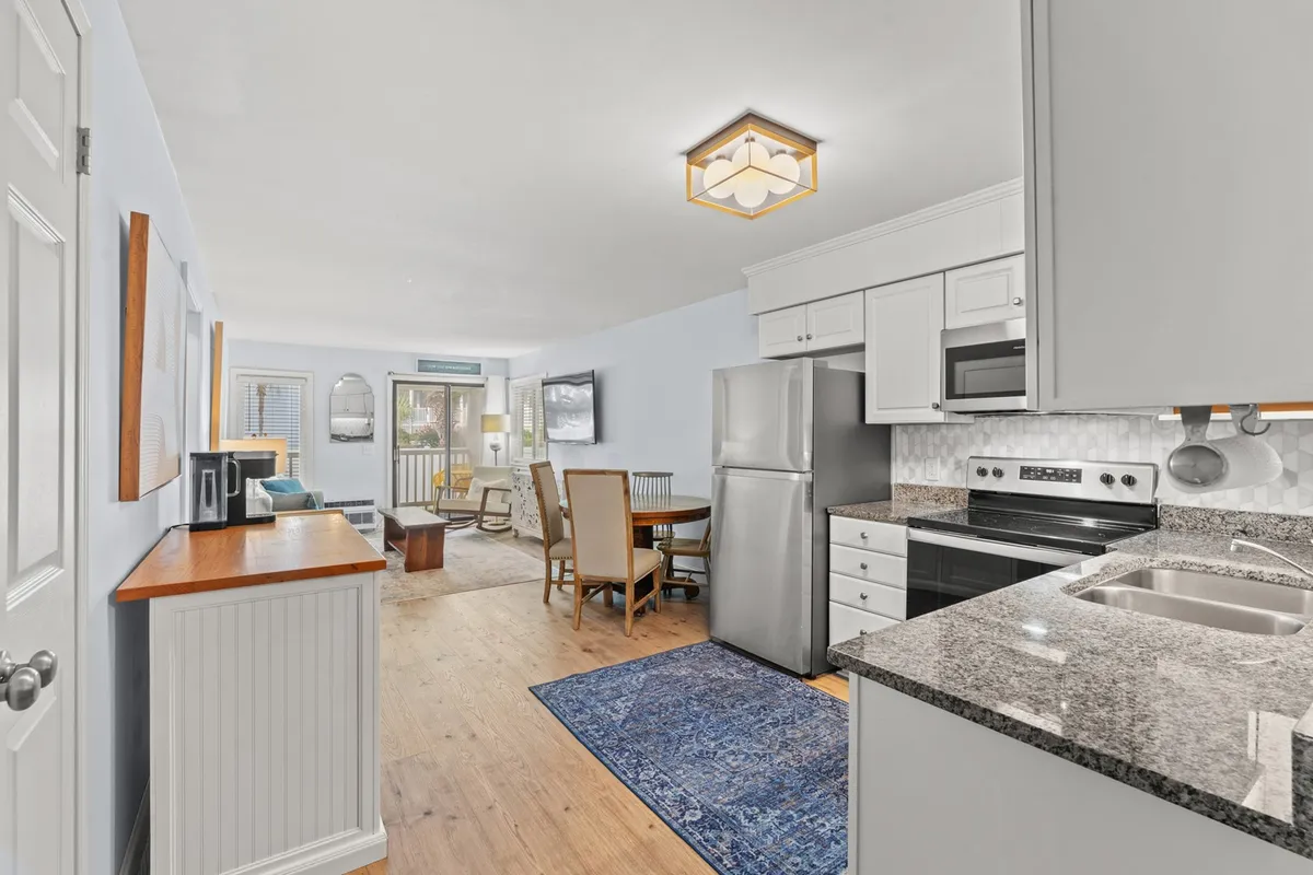 A view of the open kitchen area connected to a dining table and living space, with a granite countertop and a blue area rug tying the spaces together. The scene feels inviting and designed for shared experiences—ideal for cooking and dining together.