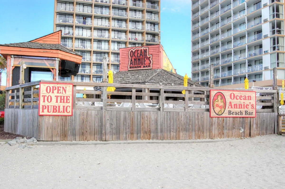 A cheerful beach bar scene with yellow umbrellas, a welcoming sign for Ocean Annie's Beach Bar, and people lounging nearby. The setting is festive and relaxed, with ocean views in the background and an inviting space to gather after a day of adventure.