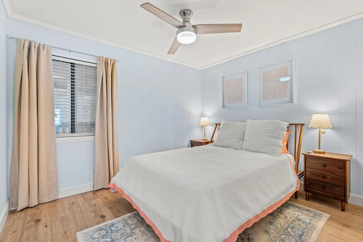 A bright, tranquil bedroom featuring a plush bed, matching nightstands, lamps, a large window, and soothing artwork. Sunlight filters in, emphasizing the airy space ideal for unwinding after a busy day or enjoying a quiet evening.