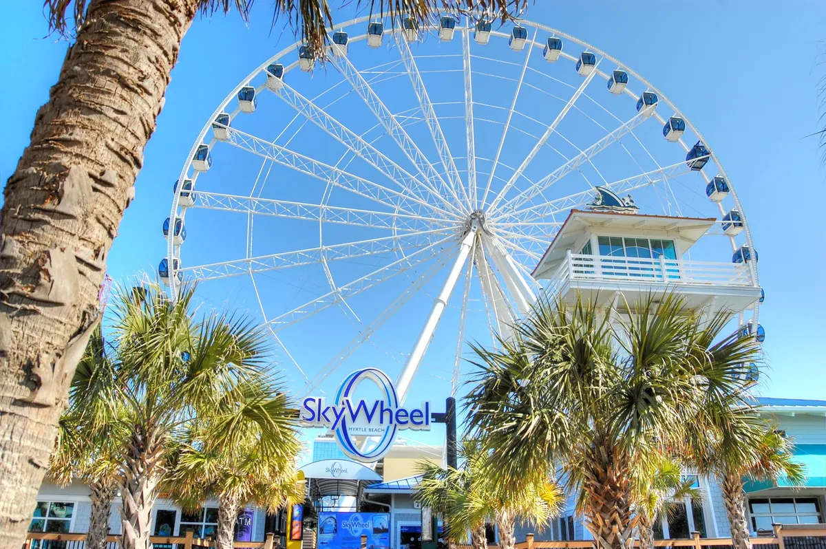 The iconic SkyWheel rising above palm trees, with a ticket booth nearby and a clear view of the ride’s colorful cabins. People can be seen lining up and snapping photos, capturing the excitement of this famous Myrtle Beach attraction at sunset.