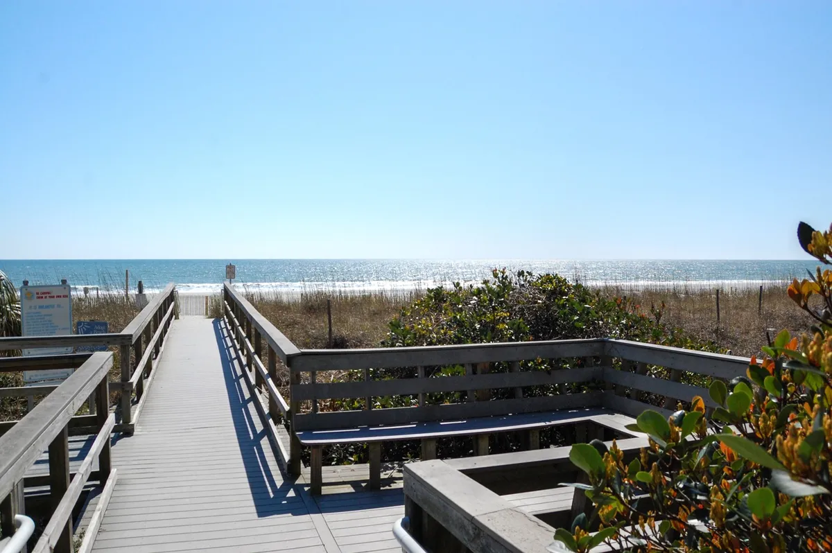A wooden boardwalk stretches through sea oats and dunes, leading directly to a wide expanse of sandy beach and the shimmering Atlantic Ocean. Gentle waves lap the shore under a soft, relaxing light, with the soothing sound of the ocean implied.