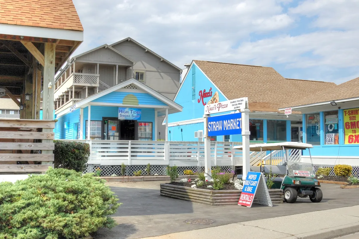 A cheerful street scene featuring the colorful facades and signage of the Straw Market and High Tide Smoke Shop, with a golf cart parked nearby. The area is bustling with activity, inviting visitors to explore shops, grab a bite, and soak in the local flavor.