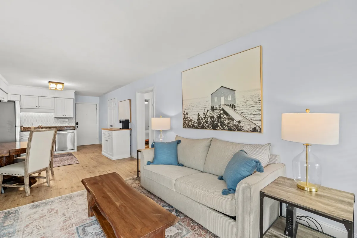 A bright, open-concept living and dining area featuring a plush sofa with blue accent pillows, a large piece of wall art depicting a house by the water, a welcoming dining table with chairs, and a lamp on a side table. Ample natural light fills the space, enhancing its cozy and inviting ambiance.