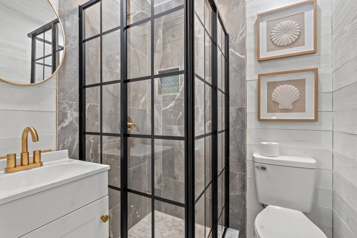 A stylish bathroom with a glass shower enclosure, a vanity with a round mirrored sink, gold fixtures, and decorative shell art on the wall. The space feels clean and refreshing, ideal for morning routines or unwinding after the day.