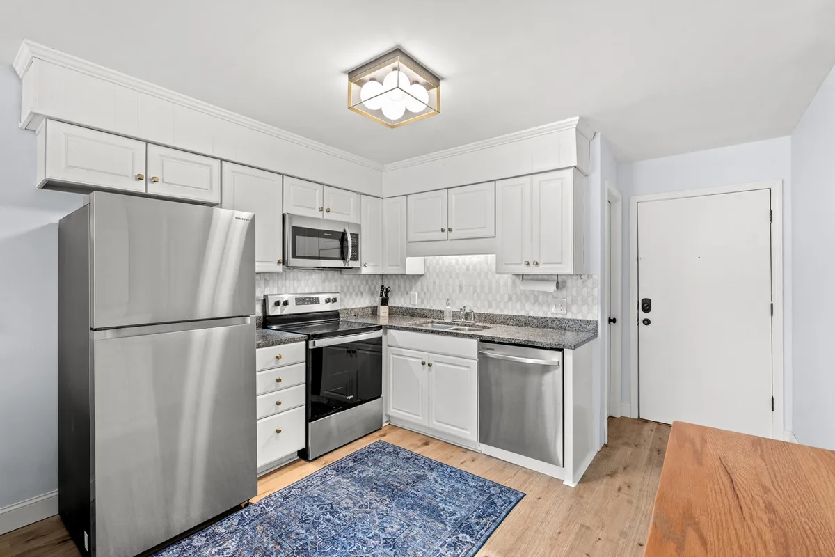 A modern kitchen featuring a stainless steel refrigerator, black stove, granite countertop, white cabinets, and a blue area rug. The space is bright, clean, and has a welcoming feel, making it ideal for both meal prep and social gatherings.