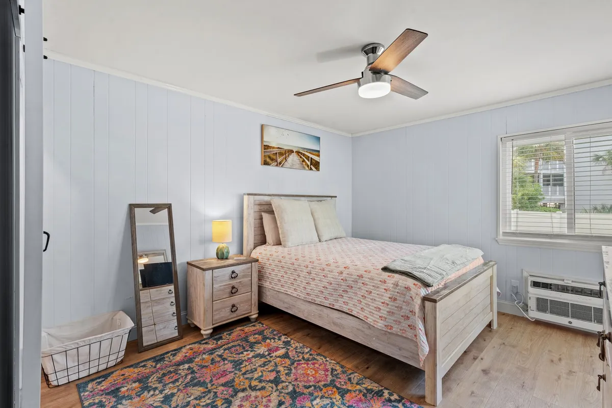 A sunlit bedroom with a queen bed, crisp linens, a soft textured rug, a nightstand, a mirror, and a ceiling fan overhead. The window lets in natural light, creating a welcoming and tranquil atmosphere perfect for relaxation or reading.