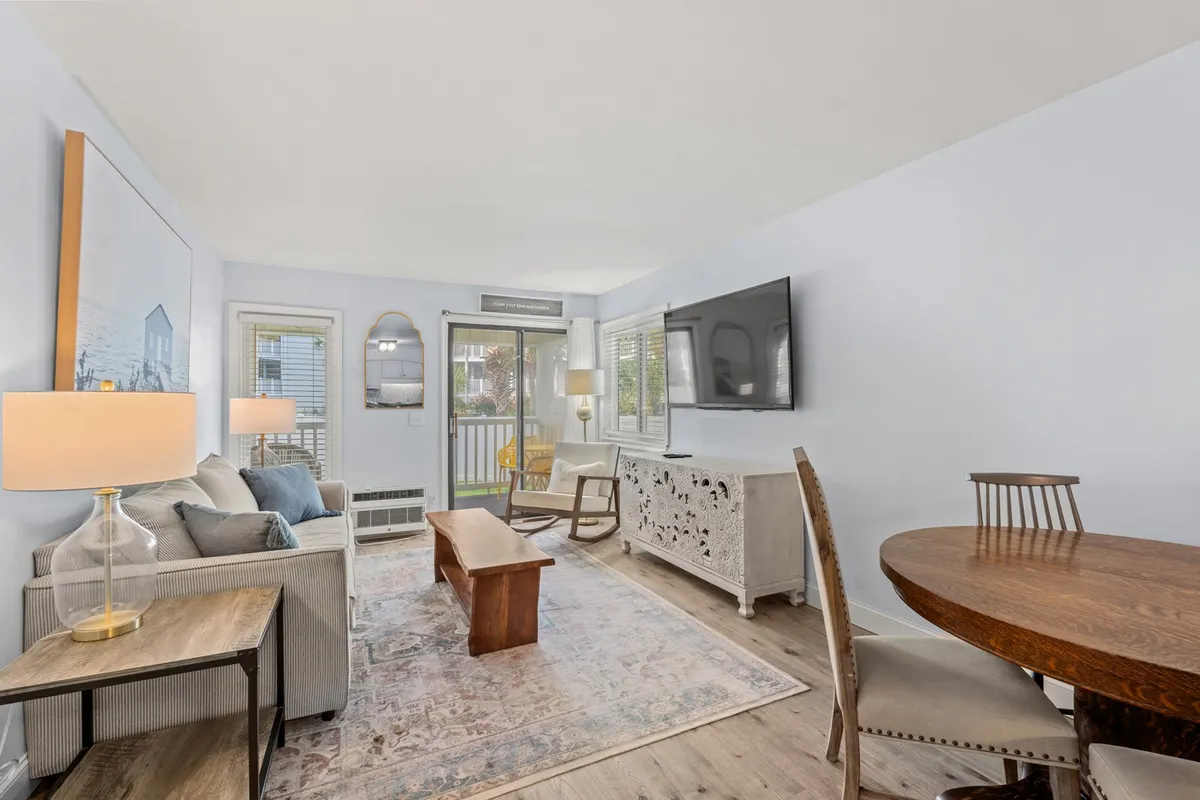 A cozy living and dining space with a plush sofa, a modern dining table, a flat-screen television, and a sliding glass door that opens onto a balcony. The room is filled with natural light, emphasizing its spaciousness and connection to the outdoors.